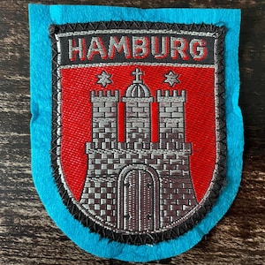 Könnte beinhalten: Ein rechteckiger gestickter Aufnäher mit einem Wappenmotiv auf hellblauem Filzhintergrund. Das Wappen zeigt eine silberne Burg auf rotem Grund und den Schriftzug HAMBURG in silbernen Buchstaben auf einem schwarzen Balken, schwarz umrandet.