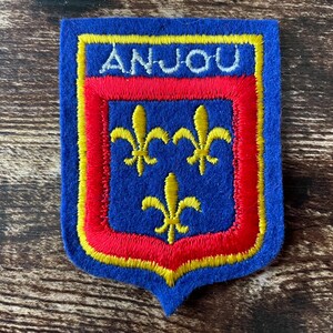 May include: A blue embroidered patch with the word "ANJOU" in white. The patch features a red and yellow border with three yellow fleur-de-lis symbols on a blue background. The patch is shield-shaped.