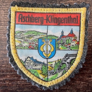 Op de afbeelding: Een schildvormige geborduurde patch met een donkergrijze vilten rand en een gele gestikte afwerking. De bovenste rode banner bevat de tekst Aschberg-Klingenthal. Het midden toont een blauw schild met een gele lier, omringd door landschapsillustraties.