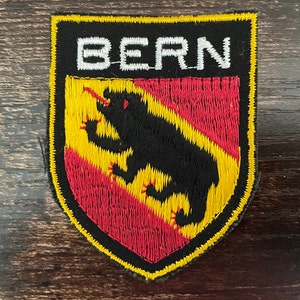 May include: Embroidered patch featuring the word "BERN" above a shield design. The shield displays a black bear over red and yellow stripes. The patch has a black border and is likely for clothing or accessories.