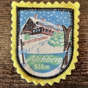 Peut inclure: Un écusson en tissu rectangulaire avec une bordure en feutrine jaune festonnée. Le centre présente une scène hivernale imprimée avec un chalet marron sous la neige, des sapins et des skis. Le texte "Nehberg 936m" est écrit en blanc sur fond bleu.