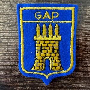 May include: A blue and yellow embroidered patch featuring the word "GAP" above a yellow castle design. The patch has a blue background with a yellow border and is likely an emblem or logo.