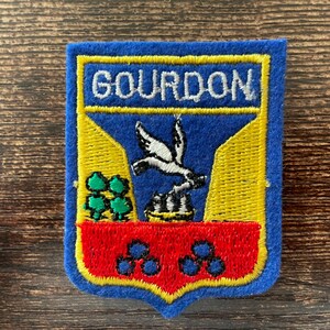 May include: A rectangular embroidered patch featuring a blue border and a shield-shaped design. The top section displays the text GOURDON in white. The center shows a white bird above a black structure, with green trees and a red base with blue circles.