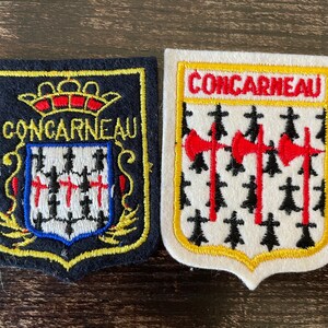 May include: Two embroidered patches featuring the text CONCARNEAU. The left patch is dark blue with a yellow border, a crown, and a shield. The right patch is white with a yellow border, featuring three red axes and black ermine spots.