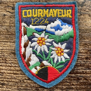 Könnte beinhalten: Ein schildförmiger gestickter Aufnäher mit dem gelben Schriftzug COURMAYEUR 1224. Das Motiv zeigt weiße Edelweißblüten mit gelben Zentren, ein blaues Gebirge, grüne Hänge, braune Felsformationen sowie eine schwarz-rote Basis.