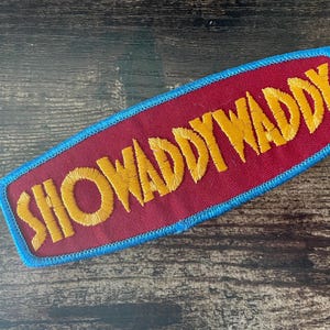 Op de afbeelding: Een rechthoekige geborduurde patch met een bordeauxrode stoffen basis en een felblauwe gestikte rand. De tekst SHOWWADDYWADDY is in vetgedrukte gele draad over het midden van de patch geborduurd in een licht schuin lettertype.