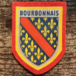 May include: A shield-shaped patch with a red felt border. The centre features a blue field with yellow fleur-de-lis symbols and a diagonal red stripe. The word BOURBONNAIS is printed in dark blue capital letters on a white background at the top.
