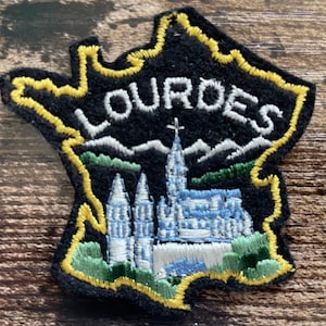 May include: A black embroidered patch shaped like the map of France with a yellow border. It features the white text LOURDES above a depiction of a light blue cathedral with spires, green foliage, and white mountains in the background.