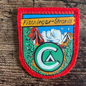 May include: A rectangular patch with a red border and a stitched edge. The patch features a colorful illustration of a campsite with the text "Fischleger-Strand" across the top. A green circle with a white "C" and a tent is in the center.