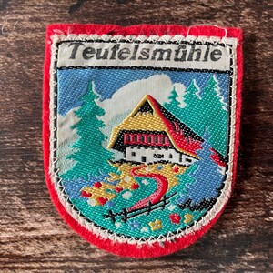 以下が含まれることがあります： 赤いフェルトの縁取りが施された盾形の刺繍ワッペン。中央には黄色い屋根の家、緑色の松の木、赤い小道が織り込まれています。上部の白い背景には、黒い文字でTeufelsmühleとプリントされています。
