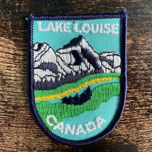 May include: A rectangular embroidered patch with rounded bottom corners featuring a light blue background, white mountains, green and yellow fields, and a dark blue border. The text LAKE LOUISE is embroidered in white at the top and CANADA at the bottom.