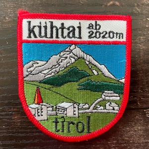 Könnte beinhalten: Gestickter Aufnäher mit rotem Rand, der eine Berglandschaft zeigt. Der Aufnäher enthält oben den Text "kuihtai ab 2020m" und unten "tirol". Das Design zeigt ein Gebirge, grüne Hügel und Gebäude.