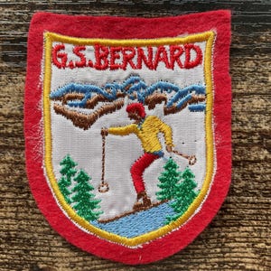 Könnte beinhalten: Ein schildförmiger, bestickter Aufnäher mit rotem Filzrand. Die Mitte zeigt einen weißen Hintergrund mit dem roten Schriftzug G.S. BERNARD über einem Skifahrer in gelber Jacke und roter Hose, blauen Bergen und grünen Tannen auf blauem Hang.