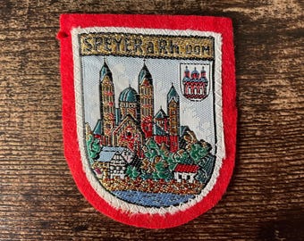 May include: A red felt patch featuring an embroidered scene of the Speyer Cathedral with the text SPEYER a.Rh. DOM. The design includes a light blue background, green trees, and a small crest with a red building on the upper right side.
