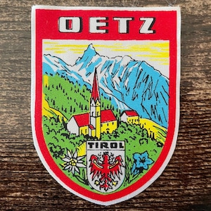 Könnte beinhalten: Ein schildförmiger gestickter Aufnäher mit dem Text OETZ oben und TIROL unten. Das Motiv zeigt eine Kirche mit rotem Dach, grüne Bäume, blaue Berge und einen gelben Himmel. Ein rotes Adler-Emblem befindet sich zentriert an der Basis.