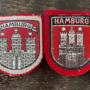 Peut inclure: Deux écussons brodés représentant les armoiries de Hambourg. Chaque écusson affiche un bouclier rouge avec un château argenté, un dôme central et le texte HAMBURG sur une bannière. L'écusson de gauche a une bordure claire, celui de droite une noire.