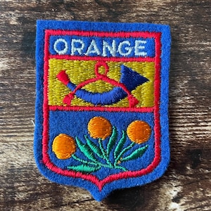 May include: Embroidered patch with a blue shield shape, featuring the word "ORANGE" at the top. The patch has a yellow and blue design in the center, with three orange flower-like shapes at the bottom, outlined in red.