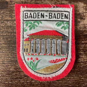 May include: A rectangular patch with a red border and a white stitched edge. The patch features the text "BADEN-BADEN" above an illustration of a building with a red roof and white columns. Green leaves and red flowers are also depicted.