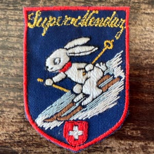 Könnte beinhalten: Ein rechteckiger gestickter Aufnäher mit blauem Hintergrund und rotem Rand. Er zeigt einen weißen Hasen beim Skifahren mit gelben Stöcken, ein kleines rotes Schild mit einem weißen Kreuz unten und den gelben kursiven Text Superallendar oben.