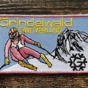 Może przedstawiać: Prostokątna haftowana naszywka na jasnoniebieskim tle z żółtym napisem Grindelwald SWITZERLAND. Przedstawia narciarza w różowym kombinezonie z żółtymi rękawicami i nartami, ośnieżoną górę oraz czarne okrągłe logo z literą G.
