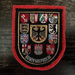 Könnte beinhalten: Gestickter Aufnäher mit schwarzem Hintergrund, rotem Rand und goldener Verzierung. Der Aufnäher zeigt mehrere farbige Wappen und die Worte "BUNDESREPUBLIK". Der Aufnäher ist wahrscheinlich ein Souvenir oder ein Emblem.