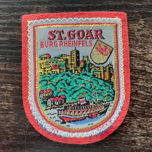 May include: A rectangular patch with a red border and a detailed embroidered design. The design features a cityscape, a boat, and the text "ST. GOAR BURG RHEINFELS". The patch has a gray background with gold, green, and red accents.