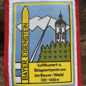 Pode incluir: Um emblema retangular bordado com borda vermelha apresentando um gráfico de um esqui amarelo, uma cordilheira branca, uma torre de igreja e pinheiros verdes. O texto diz BAYER. EISENSTEIN e Luftkurort u. Skisportzentrum im Bayer. Wald 720-1456m.