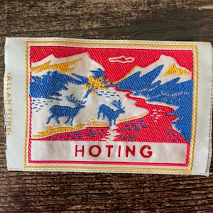 May include: A rectangular woven fabric patch featuring a landscape scene with two blue deer, white mountains, and a red sky. The word HOTING is printed in red at the bottom, and the text ALLAN FLINK is visible vertically on the left white border.