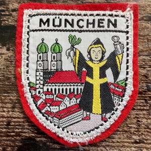 May include: A shield-shaped embroidered patch with a red felt border. The center features the text MÜNCHEN above a depiction of the Munich skyline with green-domed towers, red-roofed buildings, and a figure in a black and yellow robe.
