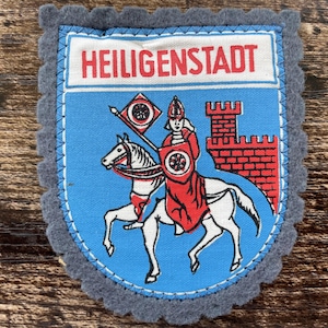 May include: A fabric patch with a scalloped gray felt border featuring a blue background, a red brick wall, and a white horse ridden by a figure in red robes holding a shield and flag. The text HEILIGENSTADT is printed in red on a white banner.