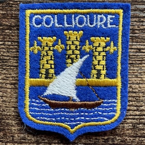 May include: A blue felt patch in a shield shape featuring yellow embroidered towers and fleur-de-lis symbols above a brown sailboat with a white sail on blue wavy lines. The word COLLIOURE is embroidered in white at the top of the patch.
