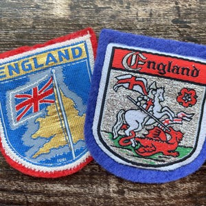 Pode incluir: Dois emblemas bordados com temas da Inglaterra. O emblema esquerdo tem uma borda vermelha, a palavra ENGLAND, uma bandeira Union Jack e um mapa dourado. O direito tem uma borda azul, a palavra England e São Jorge a derrotar um dragão.