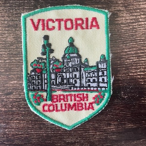 Op de afbeelding: Geborduurde patch met het woord "VICTORIA" boven een gebouwillustratie met groene koepels en de tekst "BRITISH COLUMBIA". De patch heeft een crèmekleurige achtergrond met een groene rand.