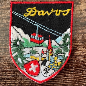 Può includere: Una toppa ricamata a forma di scudo con sfondo nero e bordo rosso. Presenta la scritta gialla Davos, una funivia rossa, montagne innevate, pini verdi, il campanile di una chiesa e due piccoli stemmi, tra cui la bandiera svizzera.