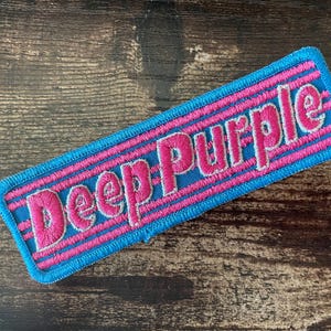 Op de afbeelding: Geborduurde patch met de woorden "Deep Purple" in roze, omlijnd met wit, op een blauwe rechthoekige achtergrond. De patch heeft roze strepen en een blauwe rand. Geplaatst op een houten oppervlak.