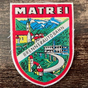 Könnte beinhalten: Ein rechteckiger Stoffaufnäher mit rotem Rand und der Aufschrift MATREI am oberen Rand. Die Abbildung zeigt eine Berglandschaft mit einer Kirche, Gebäuden und einer Autobahnbrücke, die mittig mit BRENNERAUTOBAHN beschriftet ist.