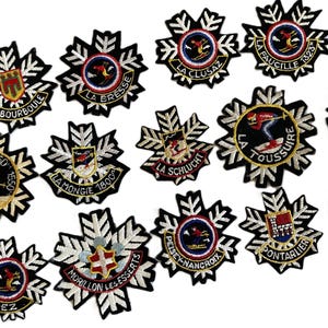 May include: A collection of embroidered patches shaped like snowflakes, each featuring a circular centre with various ski resort names and illustrations, including La Bourboule, La Bresse, La Clusaz, La Faucille, La Toussuire, La Schlucht, La Mongie, Morillon, Morez, Peisey-Nancroix, and Pontarlier.