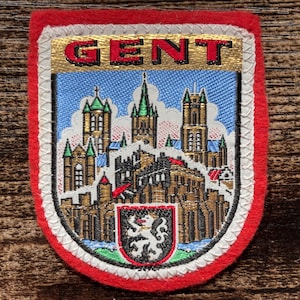 以下が含まれることがあります： 赤いフェルトの縁取りが施された長方形の刺繍ワッペン。盾形のデザインが特徴で、金色の背景に赤い文字で「GENT」と記されています。その下には、塔が立ち並ぶ都市のスカイラインと小さな紋章が詳細に刺繍されています。