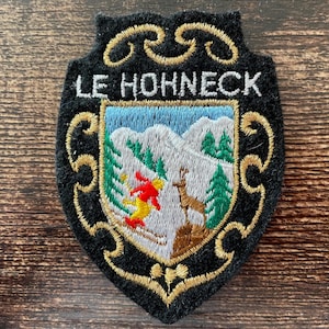 May include: A shield-shaped embroidered patch with a black background and gold scrollwork borders. The text LE HOHNECK is stitched in white. The centre features a mountain scene with green pine trees, a brown chamois, and a skier in red and yellow.