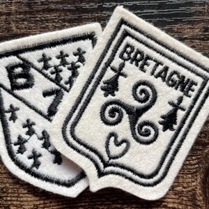 Op de afbeelding: Twee witte stoffen patches met zwart borduurwerk. Eén schildvormige patch bevat de tekst BRETAGNE, een triskelion-symbool en hermelijnvlekken. De andere patch toont de letter B, een diagonale lijn en meerdere kleine hermelijn-symbolen.