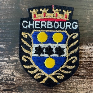 May include: Embroidered patch featuring the word "CHERBOURG" above a shield design. The shield has a blue background with yellow circles and black star-like shapes. The patch is black with gold and red accents.