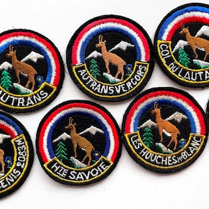 Op de afbeelding: Een verzameling ronde geborduurde patches met een bruine gems op een bergtop met groene dennenbomen en witte besneeuwde bergen. Elke patch heeft een rood, wit en blauwe rand en zwarte tekst waaronder Autrans, Autrans-Vercors, Col du Lautaret 2058M, Mt Cenis 2083M, Hte Savoie en Les Houches Mt Blanc.