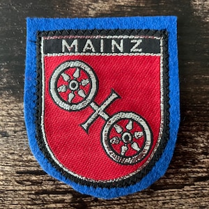 May include: A patch with a blue border and a red shield-shaped centre. The word "MAINZ" is embroidered in white on a black banner at the top. The centre features two silver-coloured wagon wheels.