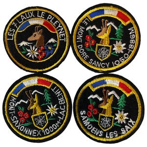 Op de afbeelding: Vier ronde geborduurde patches met zwarte randen. Elke patch toont een berggeit, een boog in de kleuren van de Franse vlag, een schild met een sneeuwvlok en tekst waaronder Les 7 Laux Le Pleynet, Le Mont Dore Sancy 1050-1886M, Mont-Saxonnex Lac Benit 1000M en Samoens Les Saix.