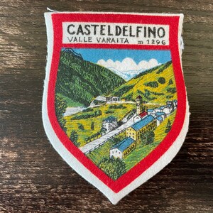 以下が含まれることがあります： ヴァッレ・ヴァライタのカスタルデルフィーノの町の風景イラストが描かれた盾形の刺繍ワッペン。上部には「CASTELDELFINO VALLE VARAITA m 1296」の文字がプリントされており、厚い赤い布の縁取りが施されています。