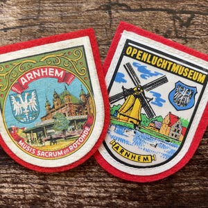 Op de afbeelding: Twee schildvormige viltemblemen met rode randen. Het linker embleem bevat de tekst ARNHEM en MUSIS SACRUM en ROTONDE met een illustratie van een gebouw en een wapenschild. Het rechter embleem toont OPENLUCHT MUSEUM en ARNHEM met een windmolen.