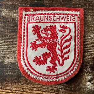 Pode incluir: Um emblema bordado retangular vermelho e branco com o texto BRAUNSCHWEIG no topo. O centro apresenta um leão vermelho sobre um fundo branco, envolvido por uma borda de escudo vermelha com costura branca ao longo das extremidades.