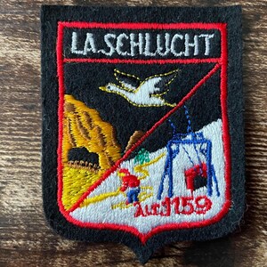 Op de afbeelding: Een rechthoekige zwarte geborduurde patch met een rode rand en de tekst LA SCHLUCHT bovenaan. Het ontwerp bevat een witte vogel, een gele rotsformatie, een skiër in rood en blauw, en een blauwe skilift met de rode tekst 1159.
