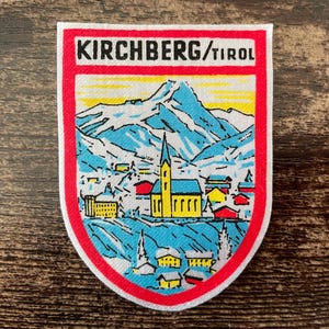 Könnte beinhalten: Ein schildförmiger Aufnäher mit einer farbenfrohen Landschaft aus Bergen und einem Dorf. Der Text KIRCHBERG/TIROL ist oben in Schwarz aufgedruckt. Das Design zeigt blaue Berge, eine gelbe Kirche sowie rote, gelbe und blaue Gebäude.