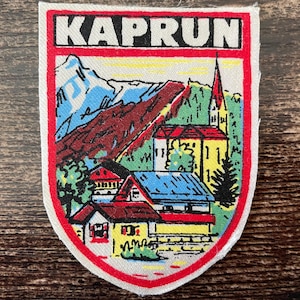 Könnte beinhalten: Ein schildförmiger gestickter Aufnäher mit dem Text KAPRUN in schwarzen Buchstaben auf weißem Grund. Das Motiv zeigt eine farbenfrohe Berglandschaft mit schneebedeckten Gipfeln, einer Kirche mit rotem Turm und mehreren Häusern mit blauen und roten Dächern, umrandet von einem dicken roten Rand.